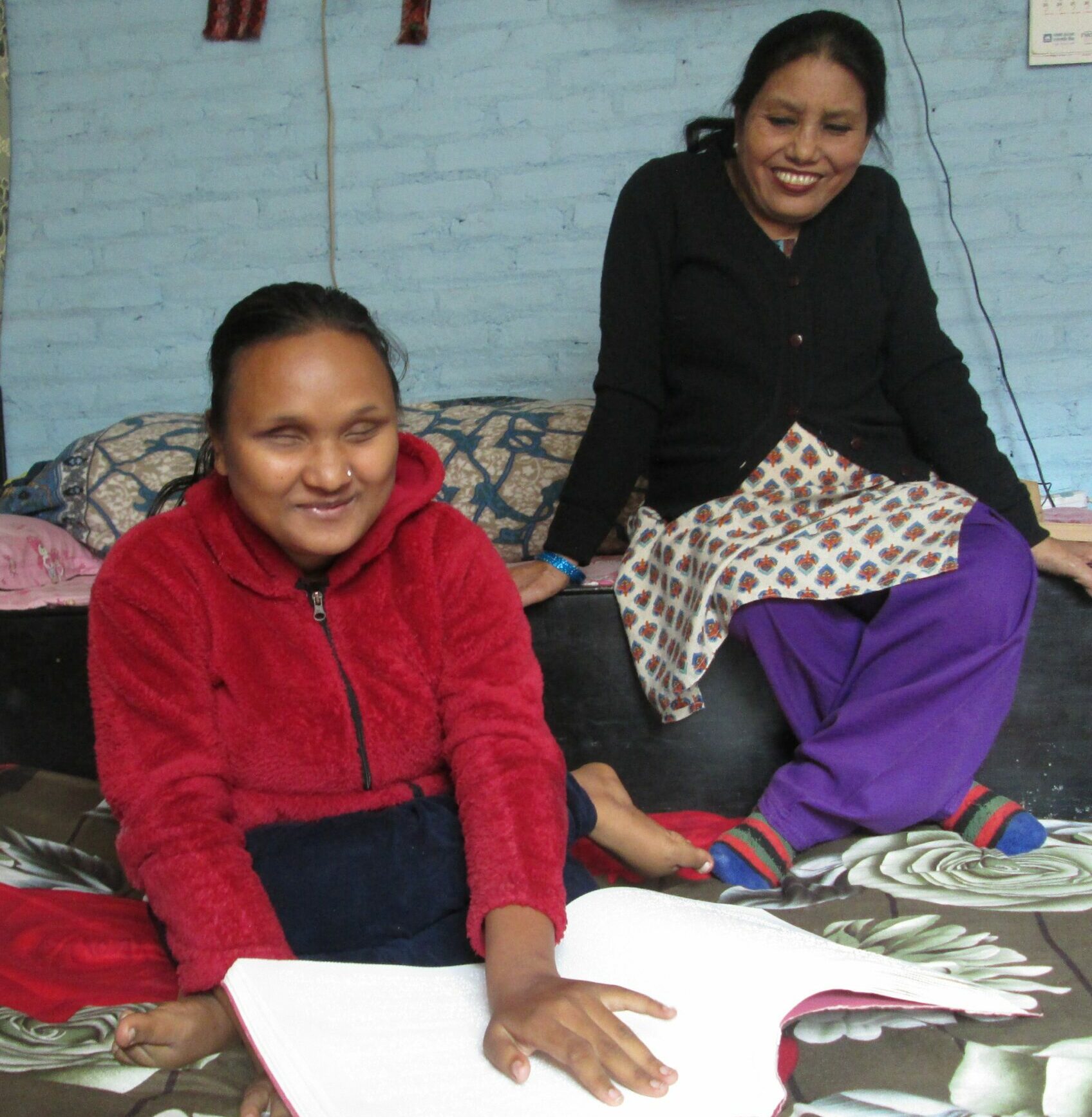 A smiling young blind woman sits on the floor with one hand on the Braille pages of a book. Her mother sits behind her, smiling.