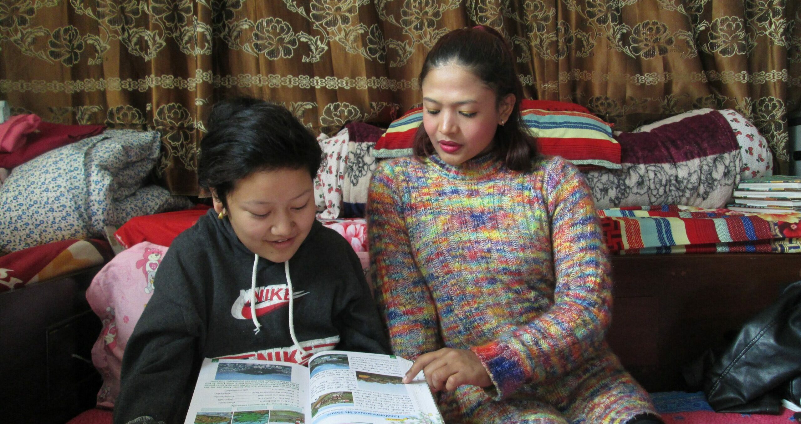 A young girl in a hoodie leans over a textbook while her tutor, a young woman, reads along beside her. Both are seated together in a homey room full of blankets and pillows.