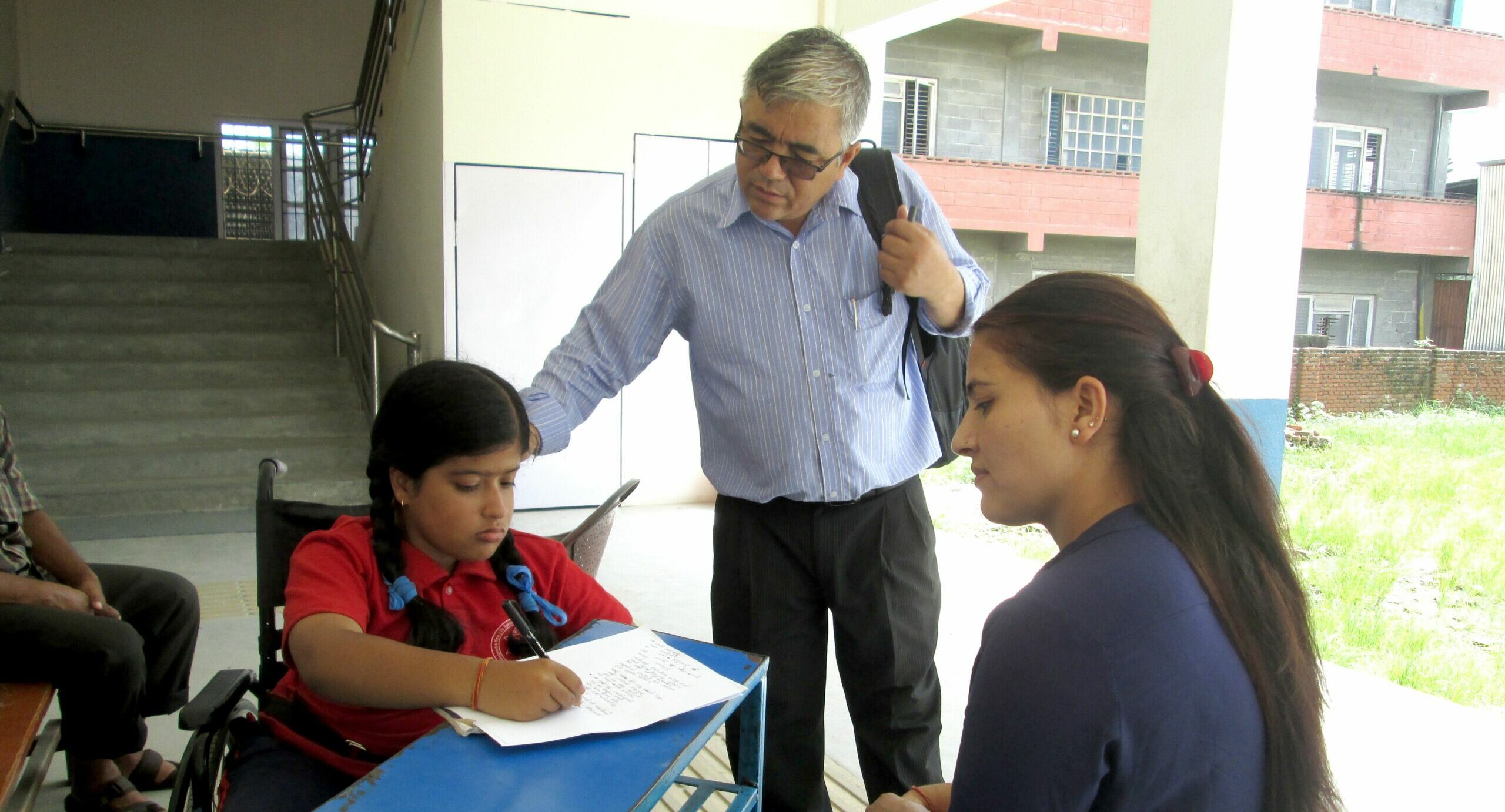 A young girl in a wheelchair works on a worksheet, seated across the table from a teacher. The girl's father stands behind, holding her backpack and placing an encouraging hand on her shoulder.