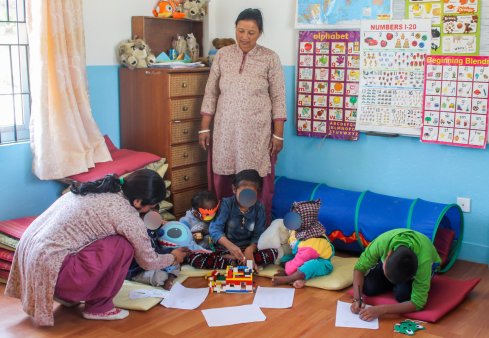 children (faces blurred to protect privacy) color together on the floor of a cheerful classroom. One adult woman looks on from above and another crouches near the children to offer feedback. the expanded mission still includes these services