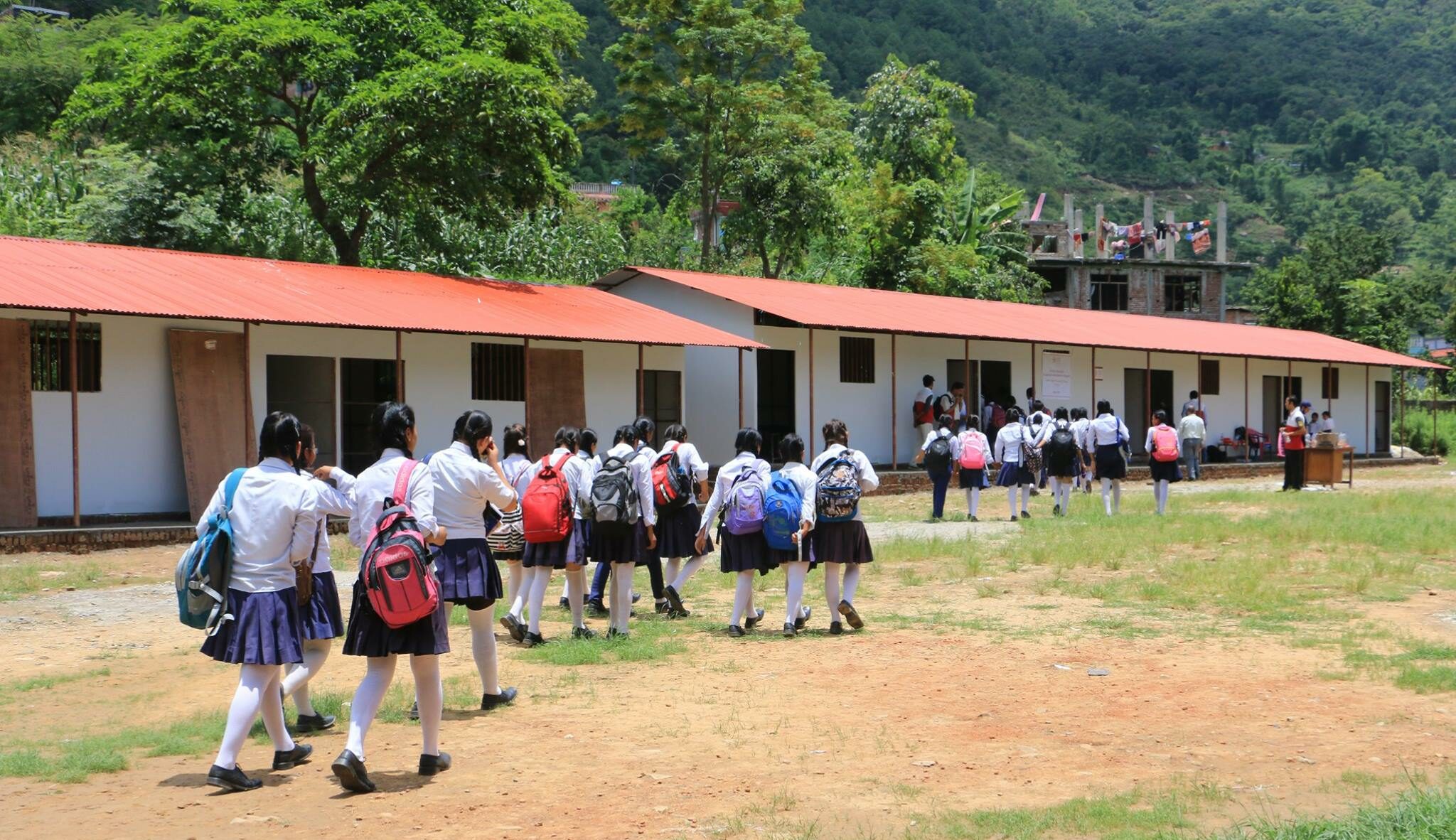 In a lush green landscape, rows of uniformed school children walk towards clean, simple classrooms with white walls and red roofs.