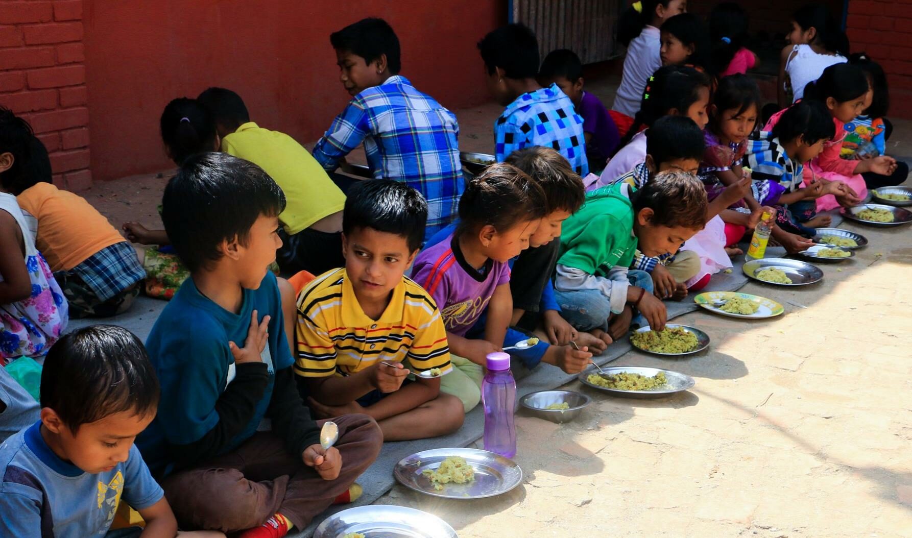 A row of children wearing colorful clothing sit in the shade eating lunch together and chatting.