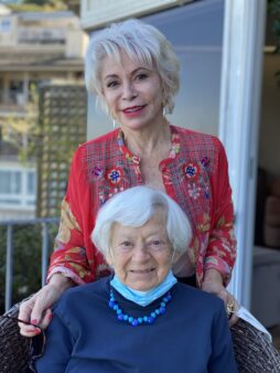 A smiling Olga Murray is seated as Medal of Freedom recipient Isabel Allende stands behind her, lovingly embracing Olga's shoulders. 