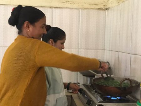 Two women stand side-by-side over a small cooktop. A woman in a green uniform instructs a woman in yellow who is stirring greens in a hot wok.