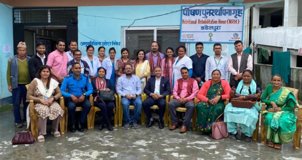 A group of 25 Nepali men and women pose in front of a blue building marked by the sign "Nutritional Rehabilitation Home"