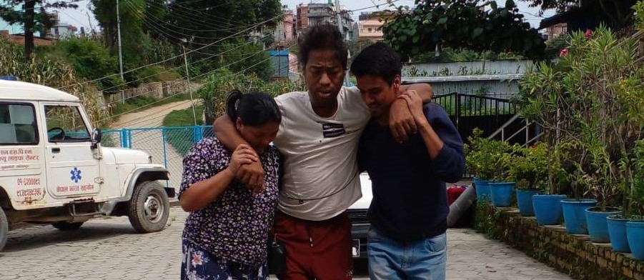 A middle-aged Nepali man walks with his weight almost entirely supported by a woman on his right side and a man on his left. Behind them, a medical truck is parked and a row of potted trees lines the sidewalk.