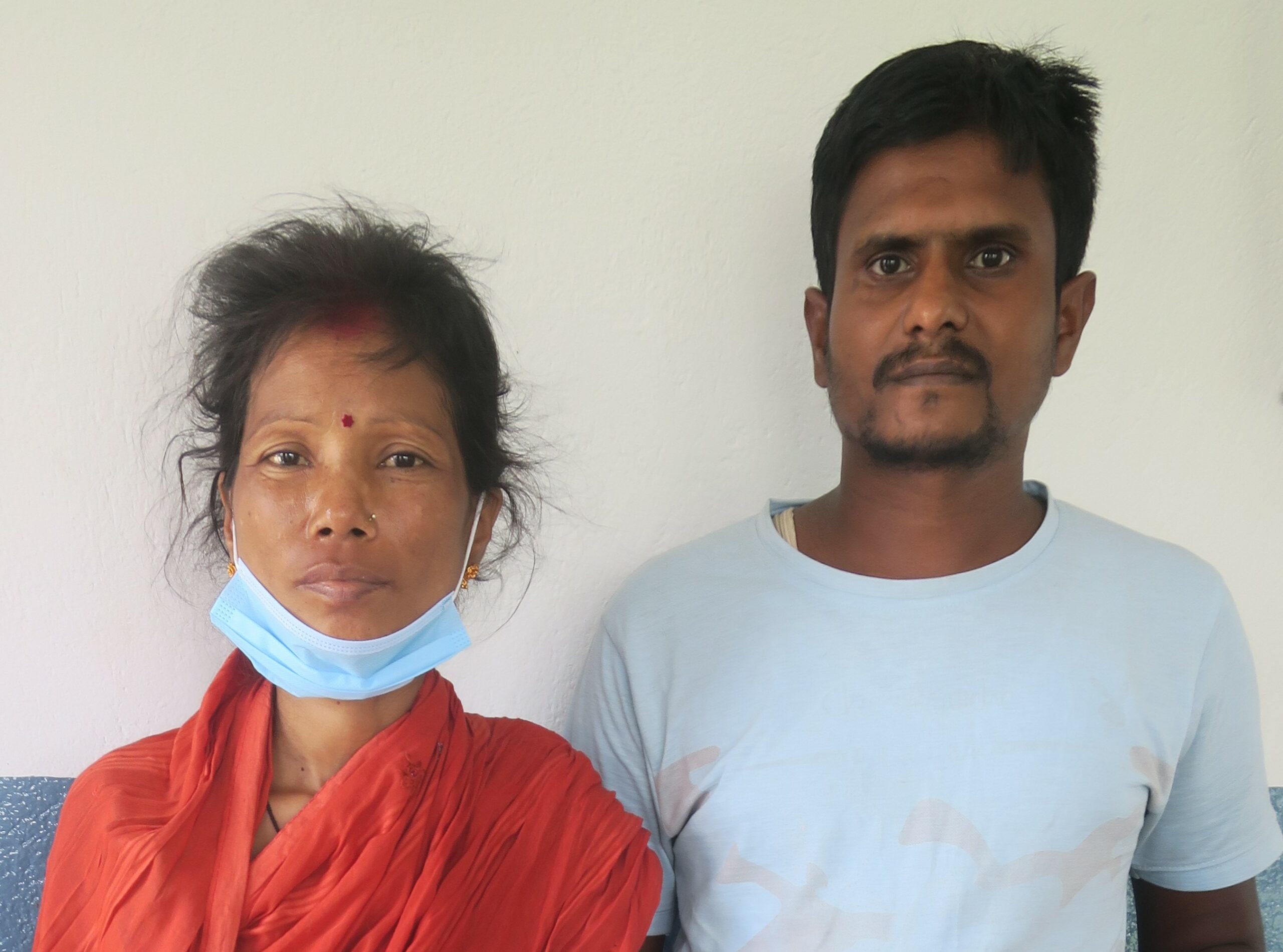 a Nepali couple in their 30s gaze calmly at the camera, looking tired. the woman is quite thin and wears an orange wrap. the man wears a plain white t-shirt.
